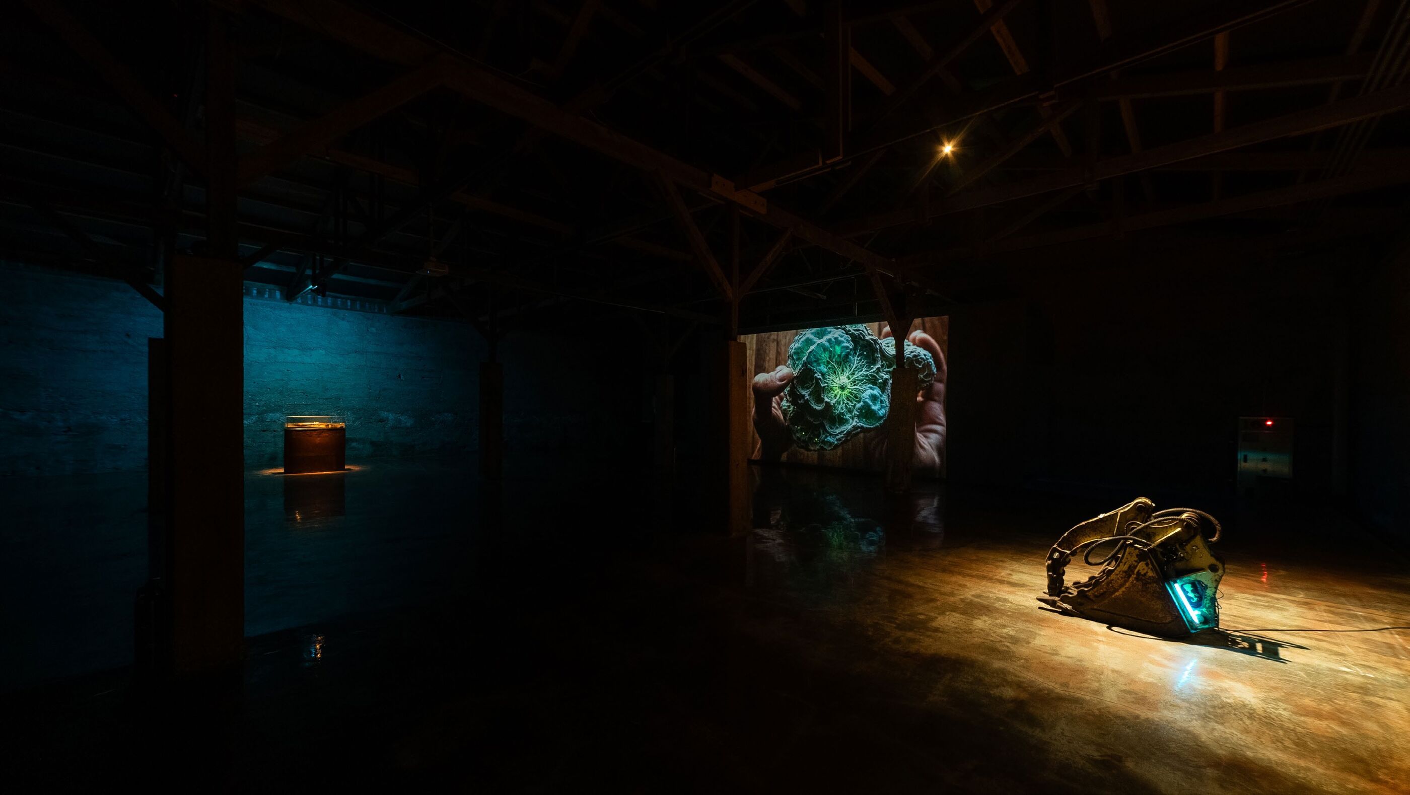 Gallery installation view: a lit glass display case on a rusted pedestal at left, a wall projection of a hand holding a glowing green resin sculpture, and a metal excavator bucket lit with blue light at right