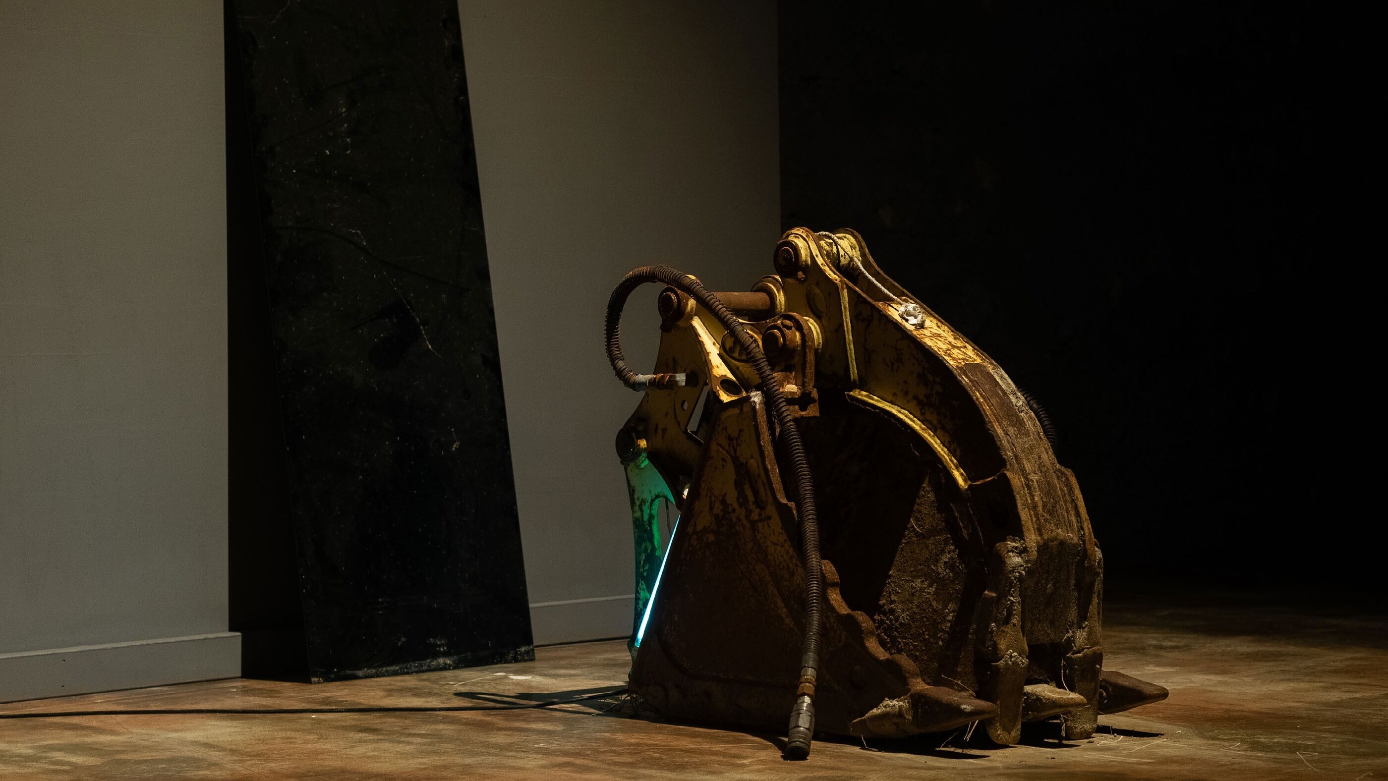 Weathered yellow excavator bucket with hydraulic hoses against a white wall and a dark angled panel, thin blue light strip visible inside the bucket