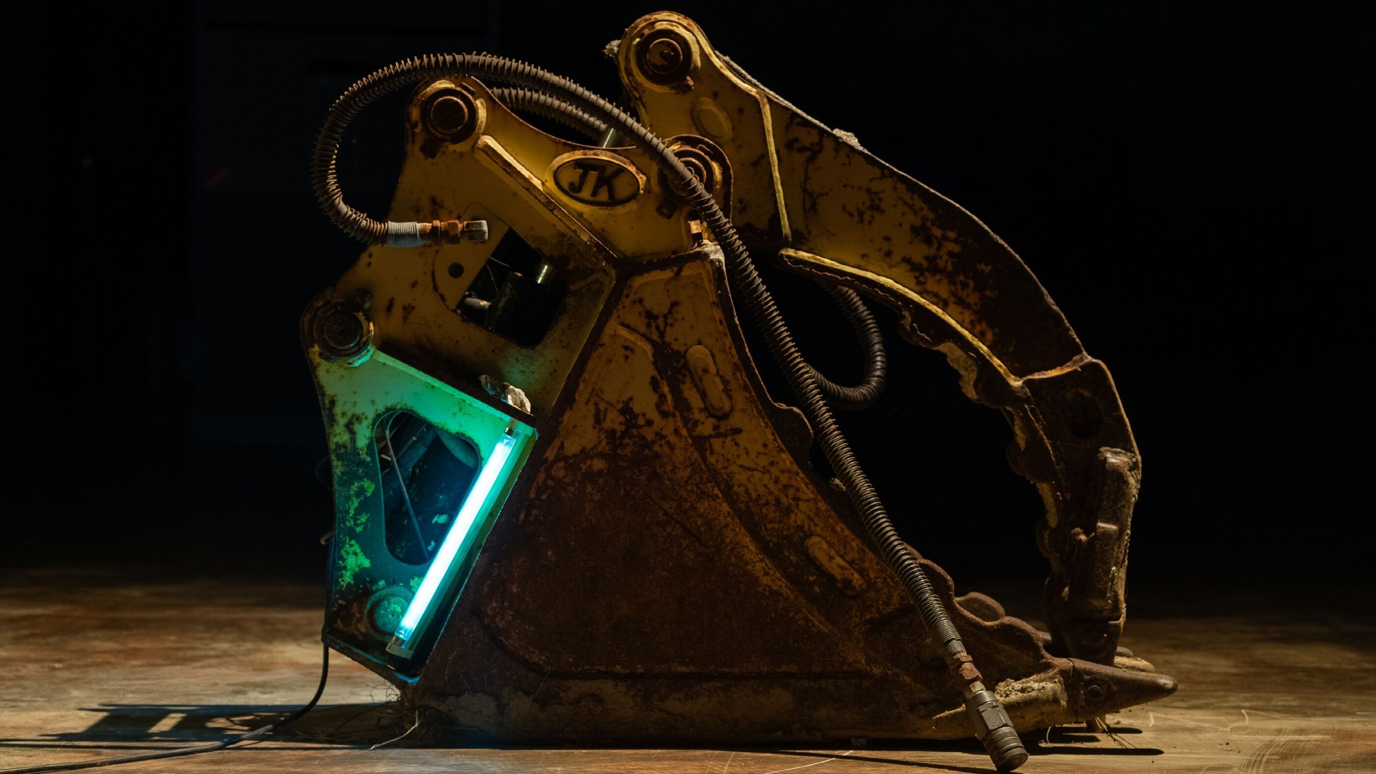 Side view of a rusted yellow excavator bucket with coiled hydraulic hoses and a thin glowing turquoise light strip inside, on a wood floor against black background