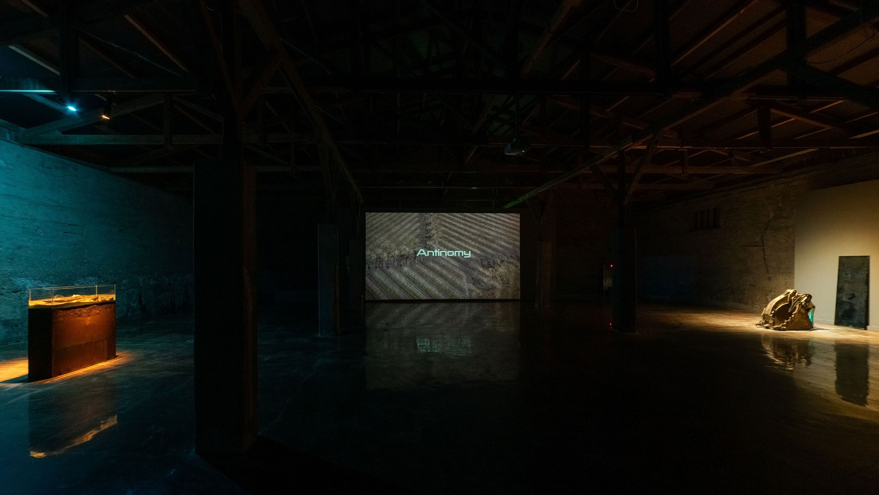 Wide dark gallery view with the word Antinomy projected on a rear wall, a lit glass case at left, and an excavator bucket sculpture lit at right
