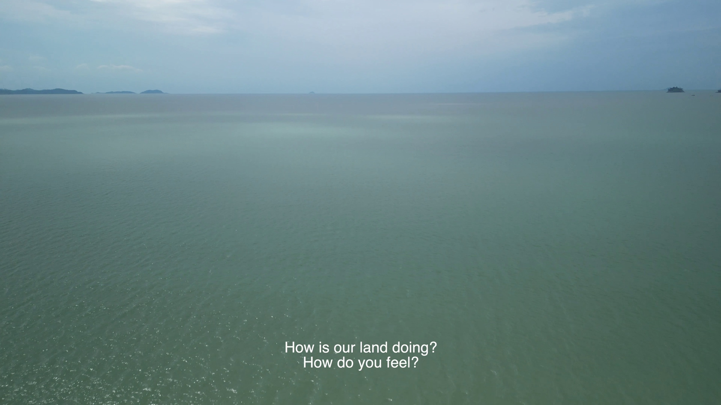 Video still of a calm sea meeting distant islands under an overcast sky, subtitle text reading 'How is our land doing? How do you feel?'