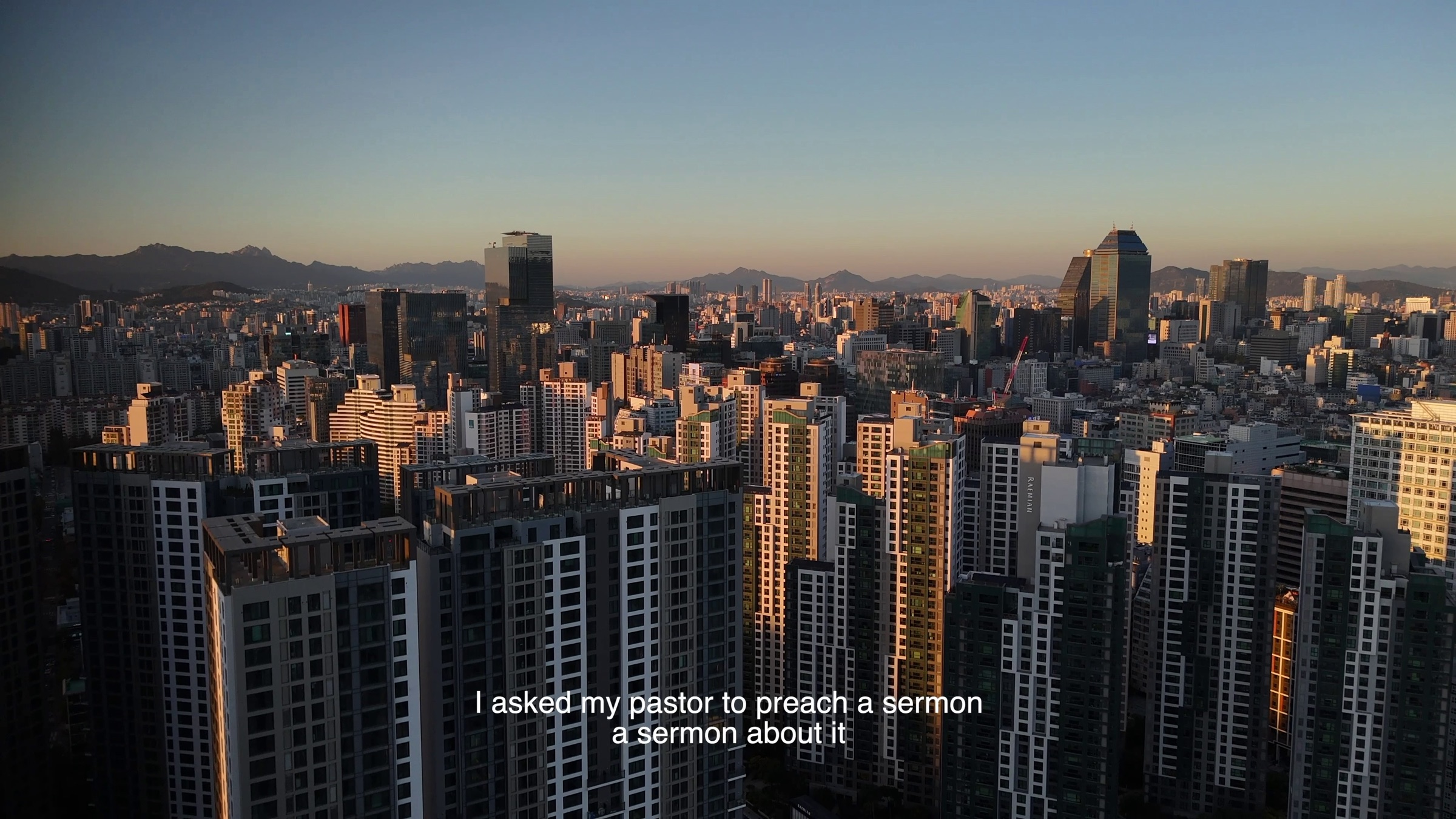 Video still of a dense high-rise city skyline at sunset with mountains behind, subtitle text reading 'I asked my pastor to preach a sermon, a sermon about it'