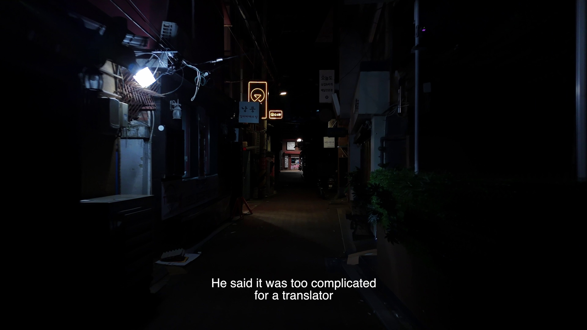Video still of a dark narrow alley at night with illuminated Korean shop signs, subtitle text reading 'He said it was too complicated for a translator'