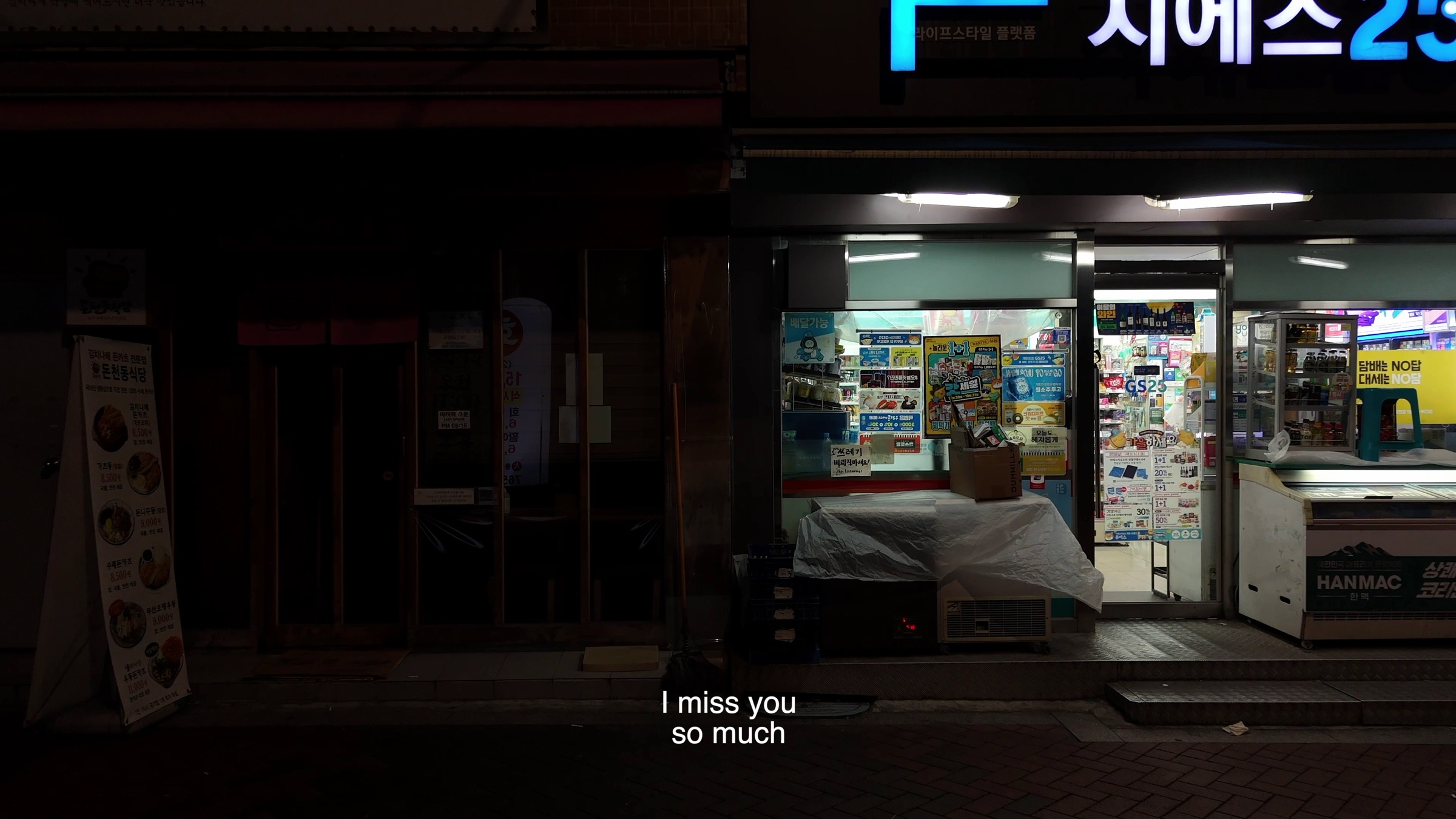 Video still of a lit convenience store front at night with posters in the window, subtitle text reading 'I miss you so much'