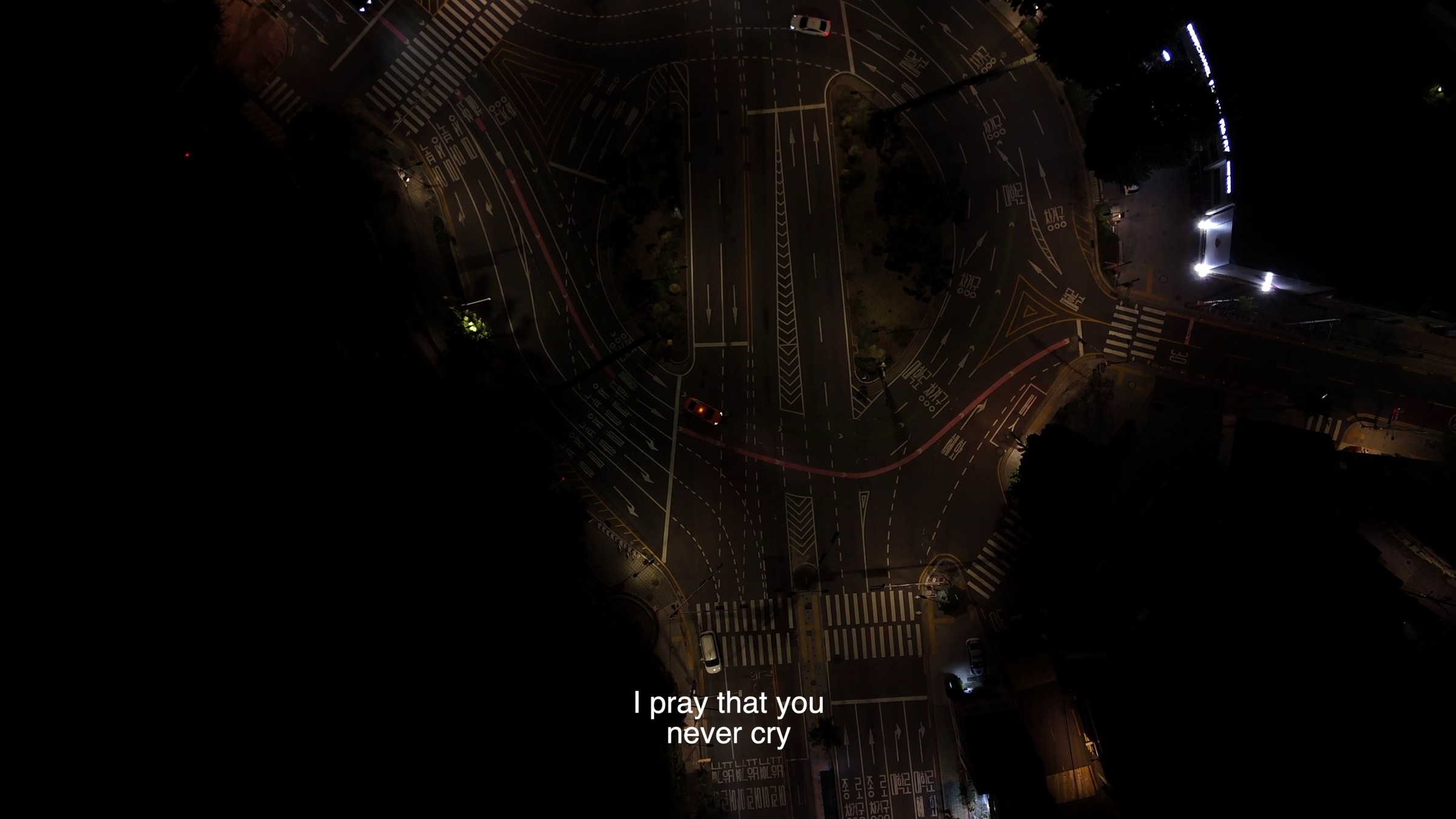 Video still of an aerial fisheye view of a night road intersection with cars, subtitle text reading 'I pray that you never cry'