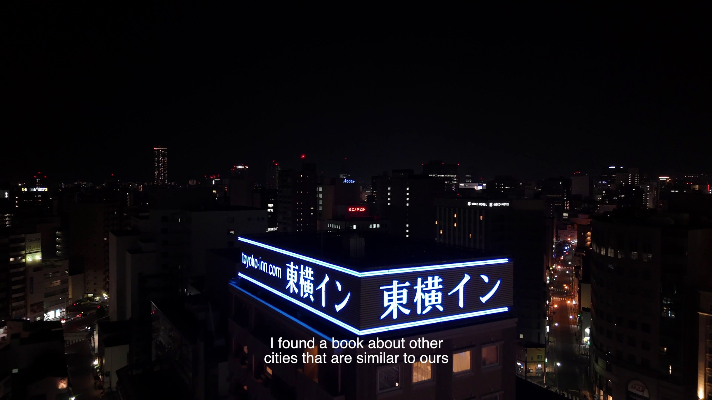 Video still of a night cityscape with an illuminated hotel rooftop sign reading Toyoko Inn, subtitle text reading 'I found a book about other cities that are similar to ours'