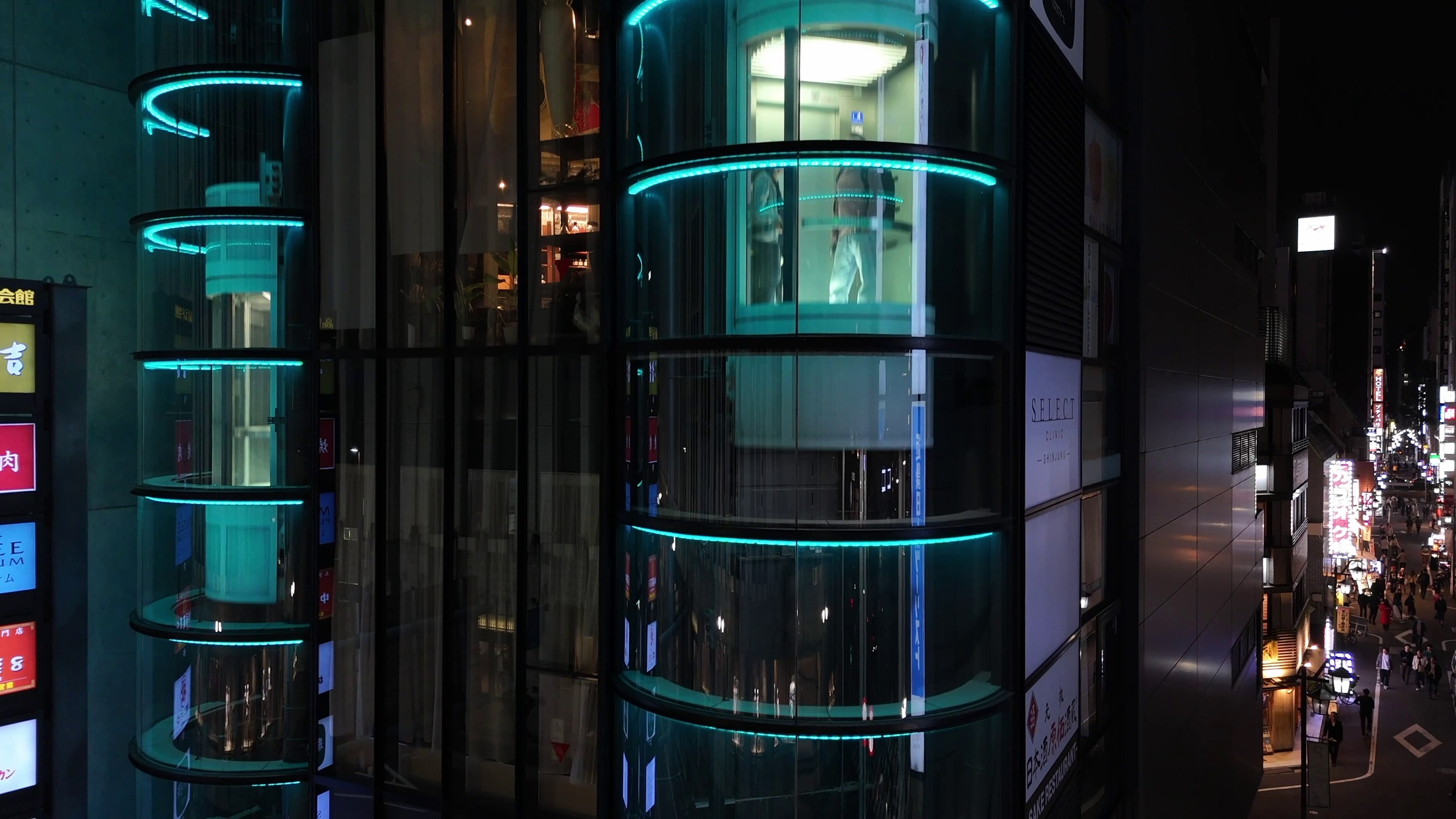 Video still of a curved glass building facade with teal neon accent lighting at night, city street with signage at right