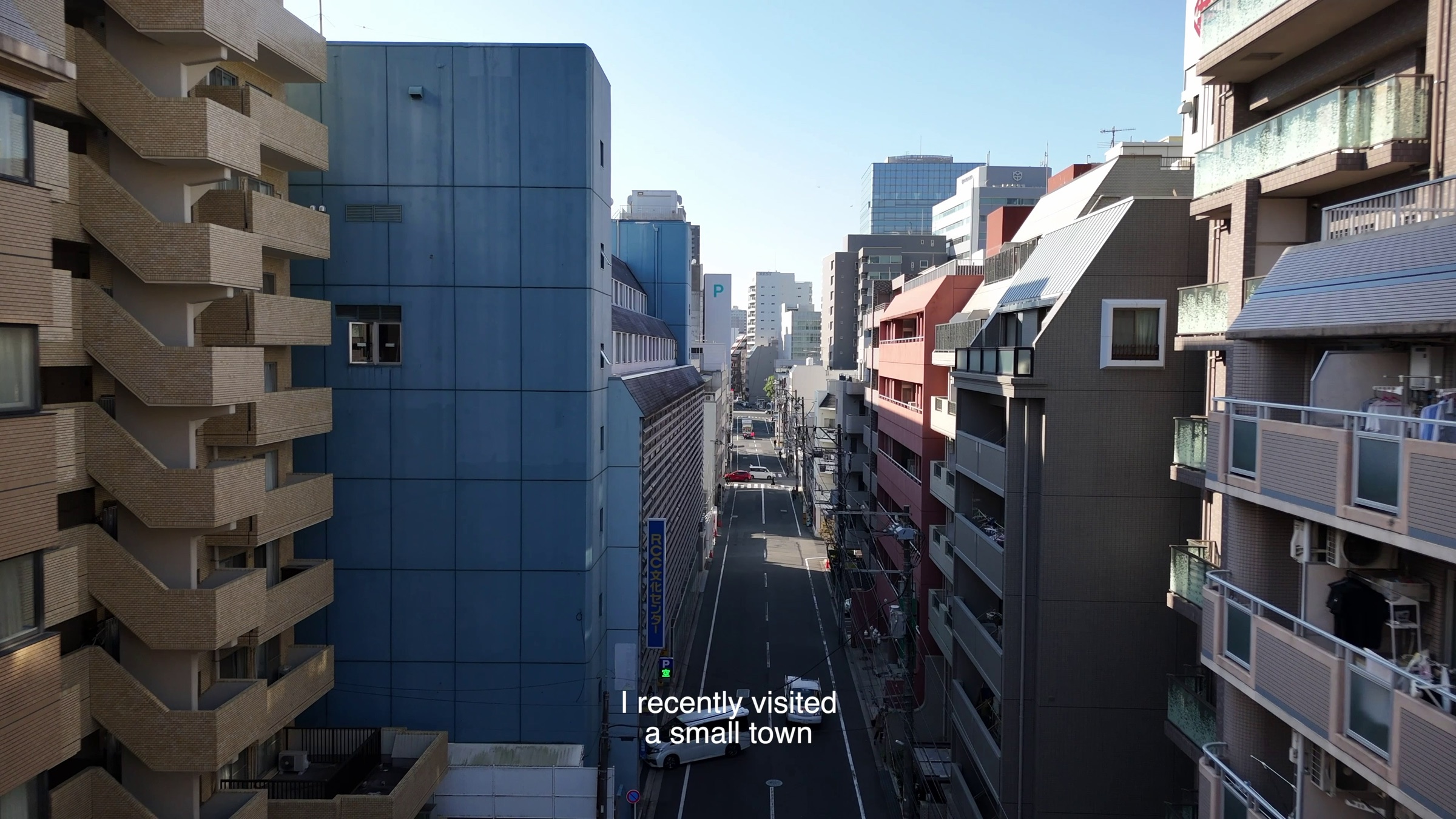 Video still of an aerial view down a street between apartment buildings under a clear sky, subtitle text reading 'I recently visited a small town'