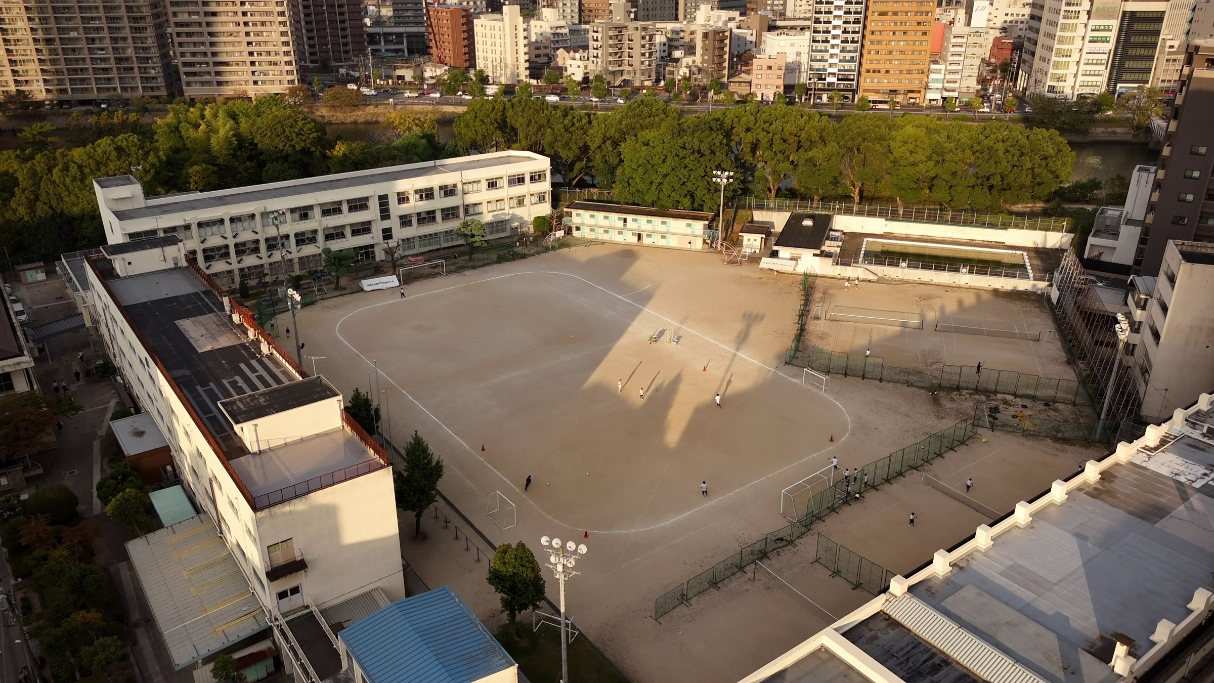 Video still of an aerial view of a school sports field with a running track, buildings, and trees along a river