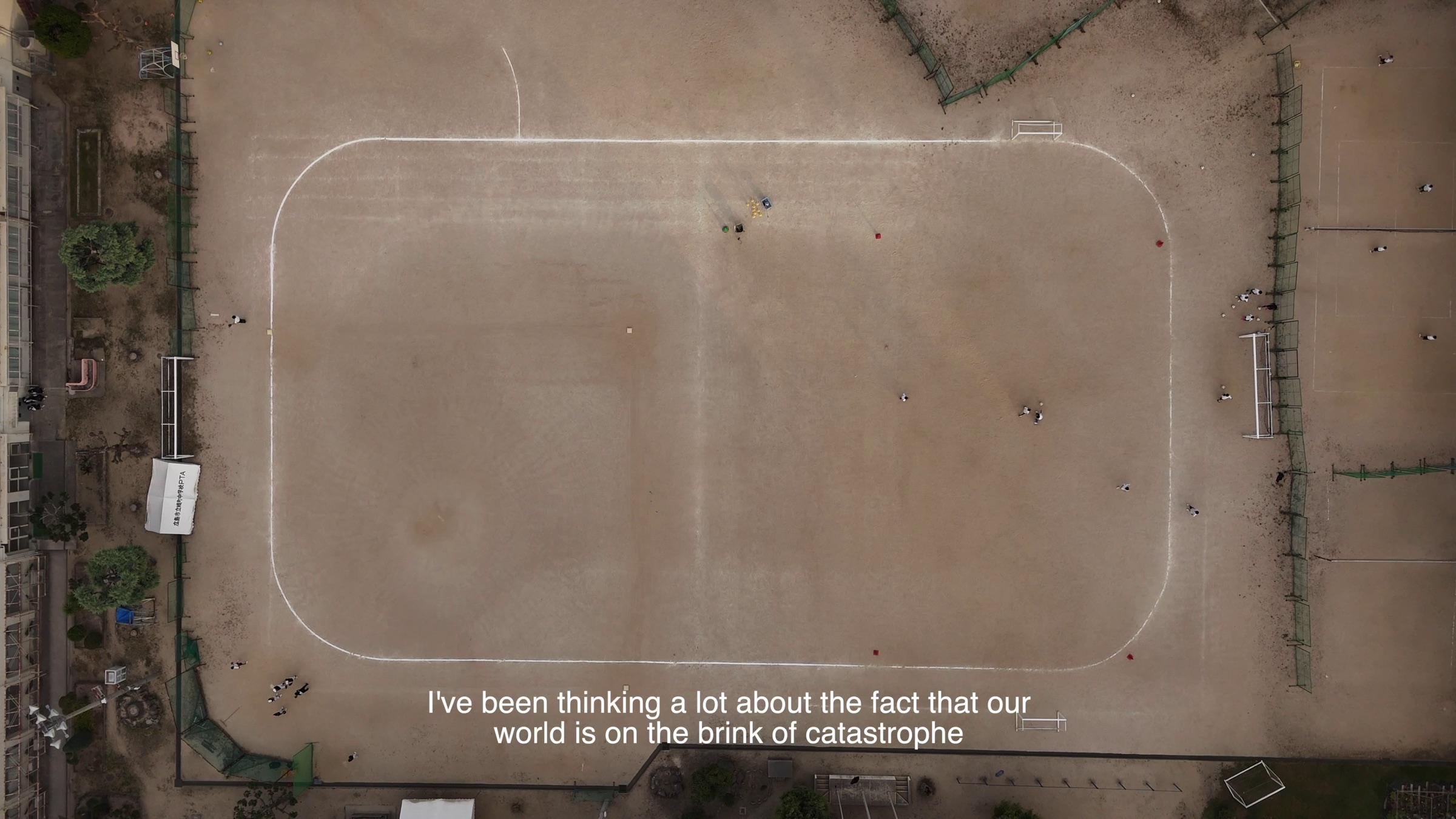 Video still of an aerial view of a school field's running track with students, subtitle text reading 'I've been thinking a lot about the fact that our world is on the brink of catastrophe'