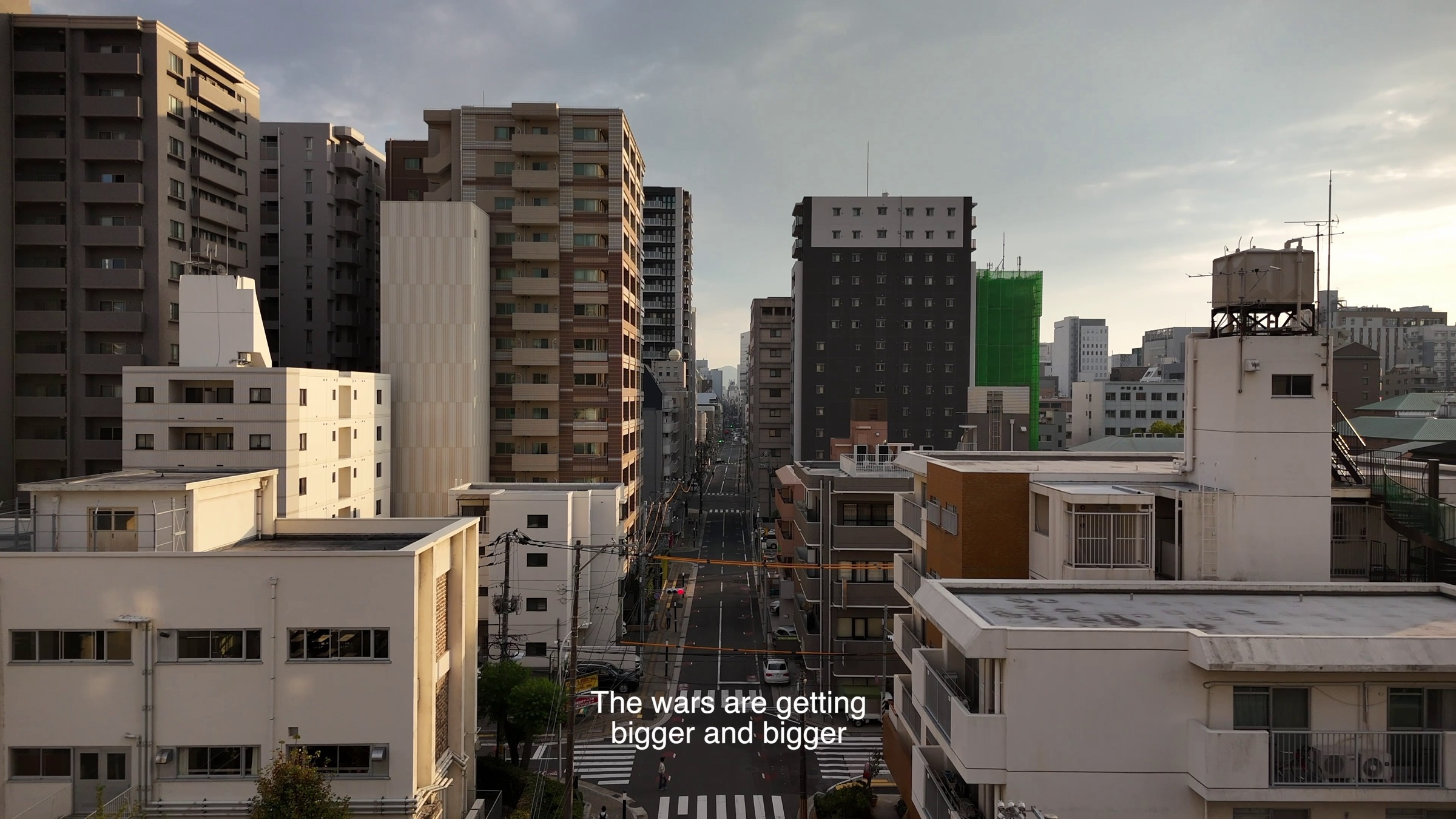 Video still of a city street seen from a rooftop between apartment buildings at dusk, subtitle text reading 'The wars are getting bigger and bigger'