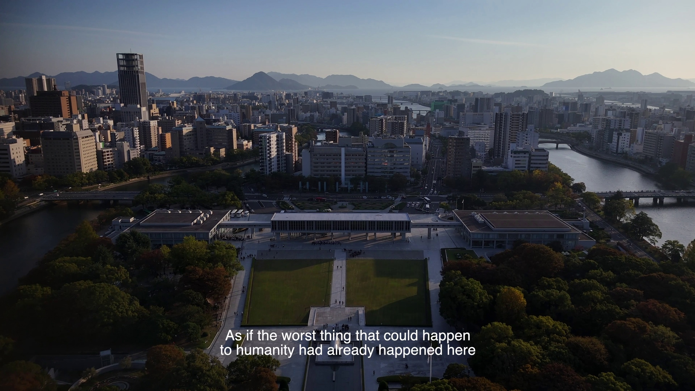 Video still of an aerial view of a memorial park with lawn and buildings beside a river and city skyline, subtitle text reading 'As if the worst thing that could happen to humanity had already happened here'