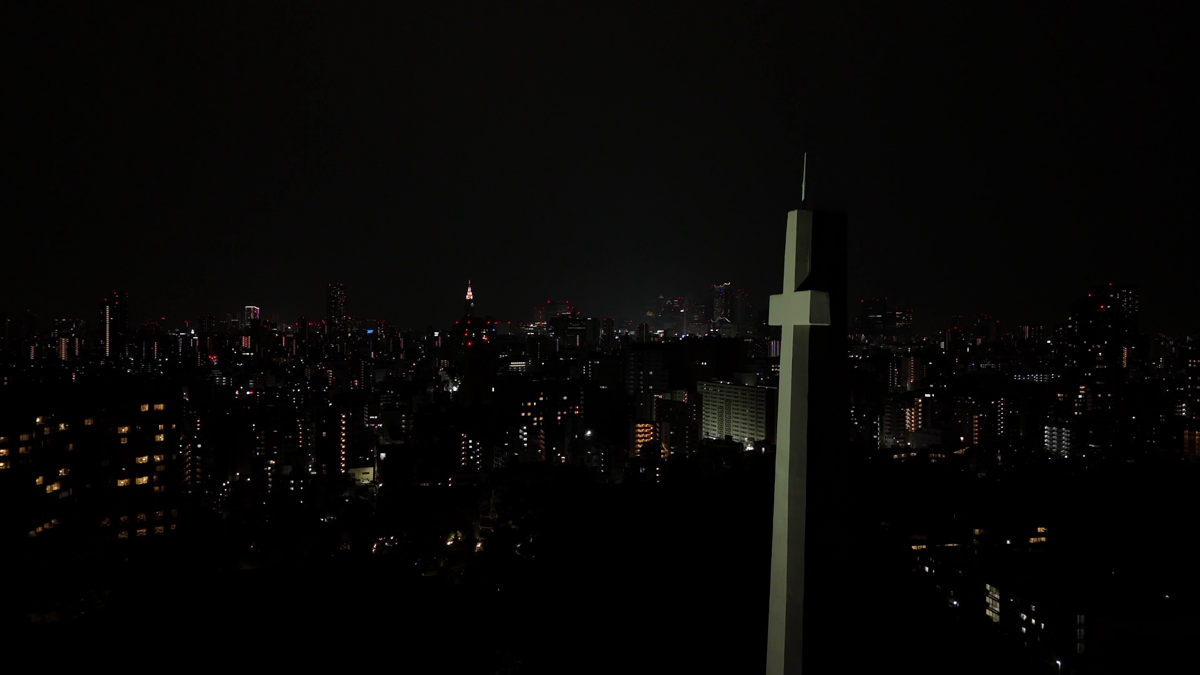 Video still of a tall illuminated cross silhouette against a dark city skyline at night
