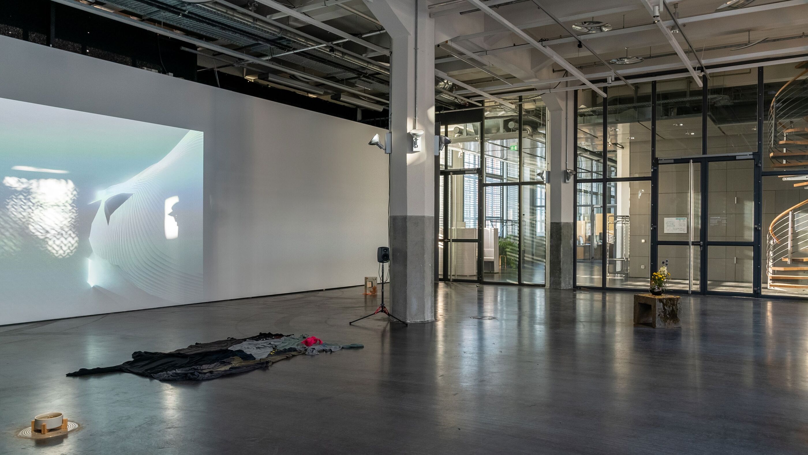 Gallery installation view with a wall projection of an abstract wavy face-like form, dark garments and a bowl on the concrete floor
