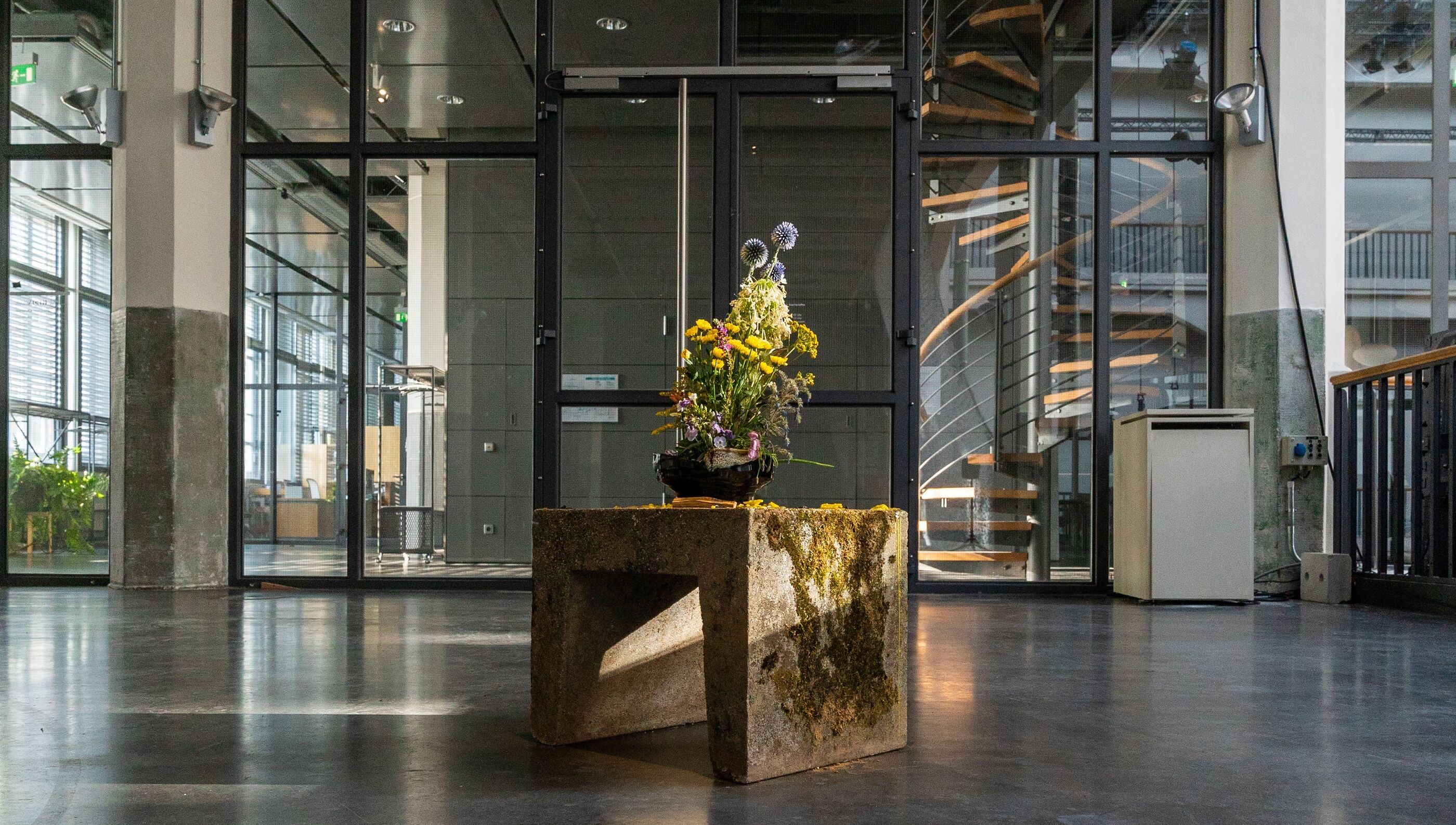 Gallery lobby view with glass doors and a spiral staircase, a floral arrangement on a moss-covered concrete plinth