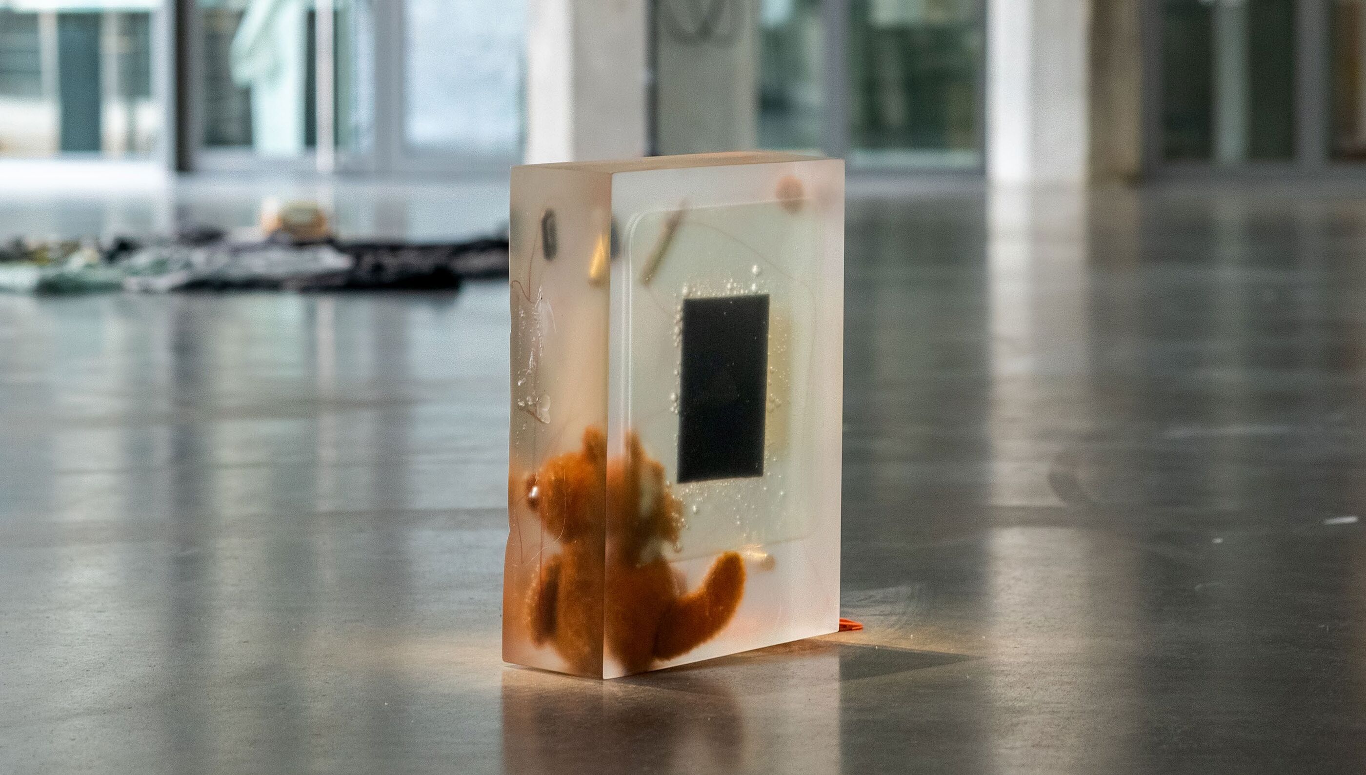 Rectangular resin block containing a small orange plush toy pressed against a dark rectangular panel, standing on a gallery floor