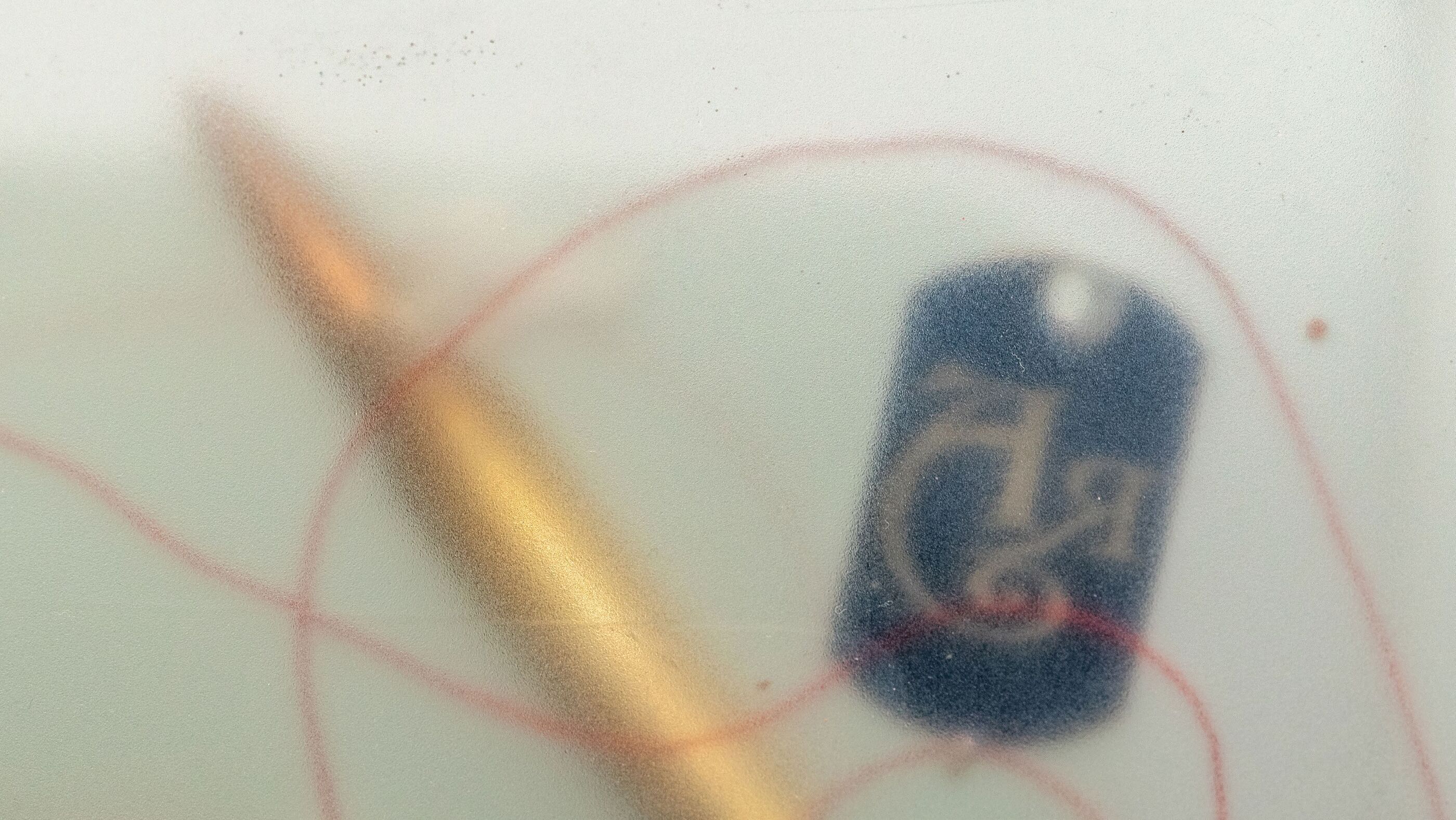 Extreme close-up through resin of a blue dog tag with partly legible lettering, red thread and a gold pin crossing in front of it, out of focus