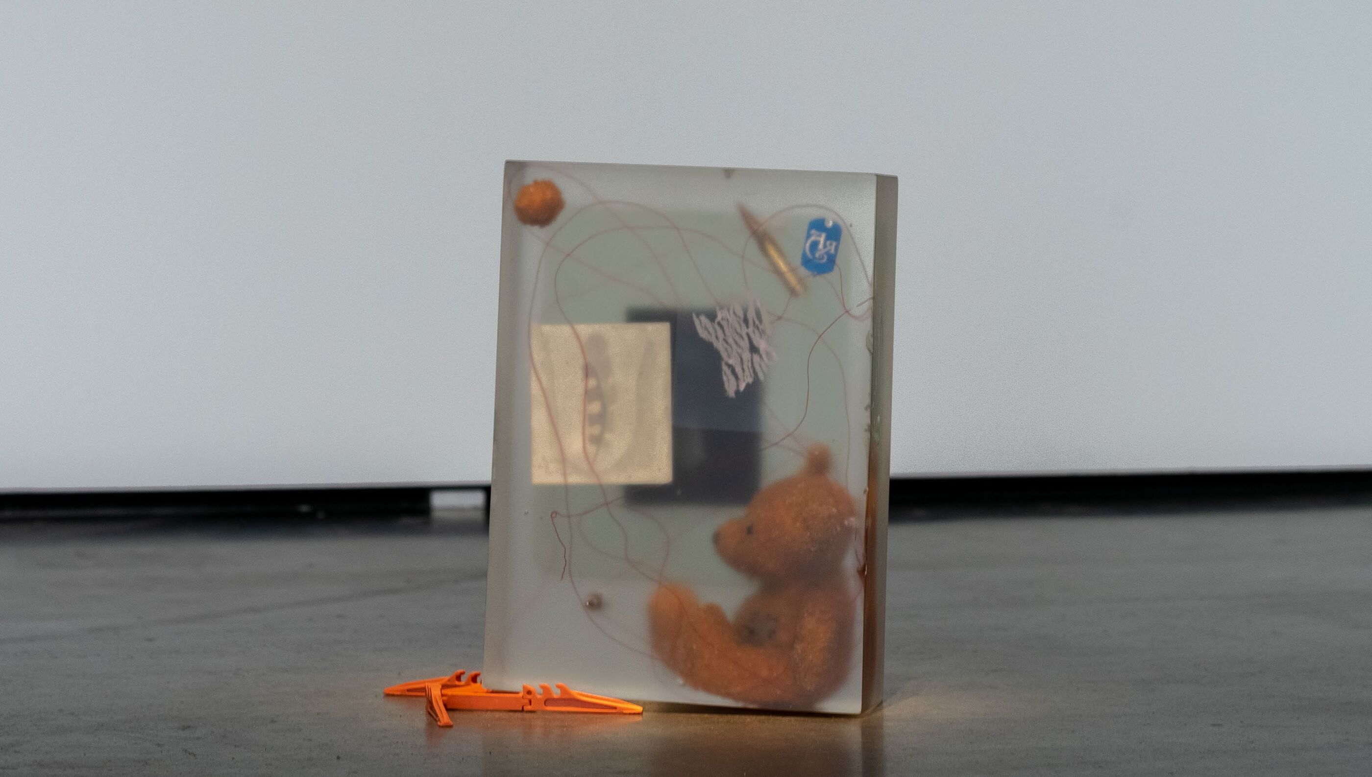 Rectangular resin block containing a small teddy bear, red thread, a bullet casing, and a blue tag, with an orange toy piece resting against its base