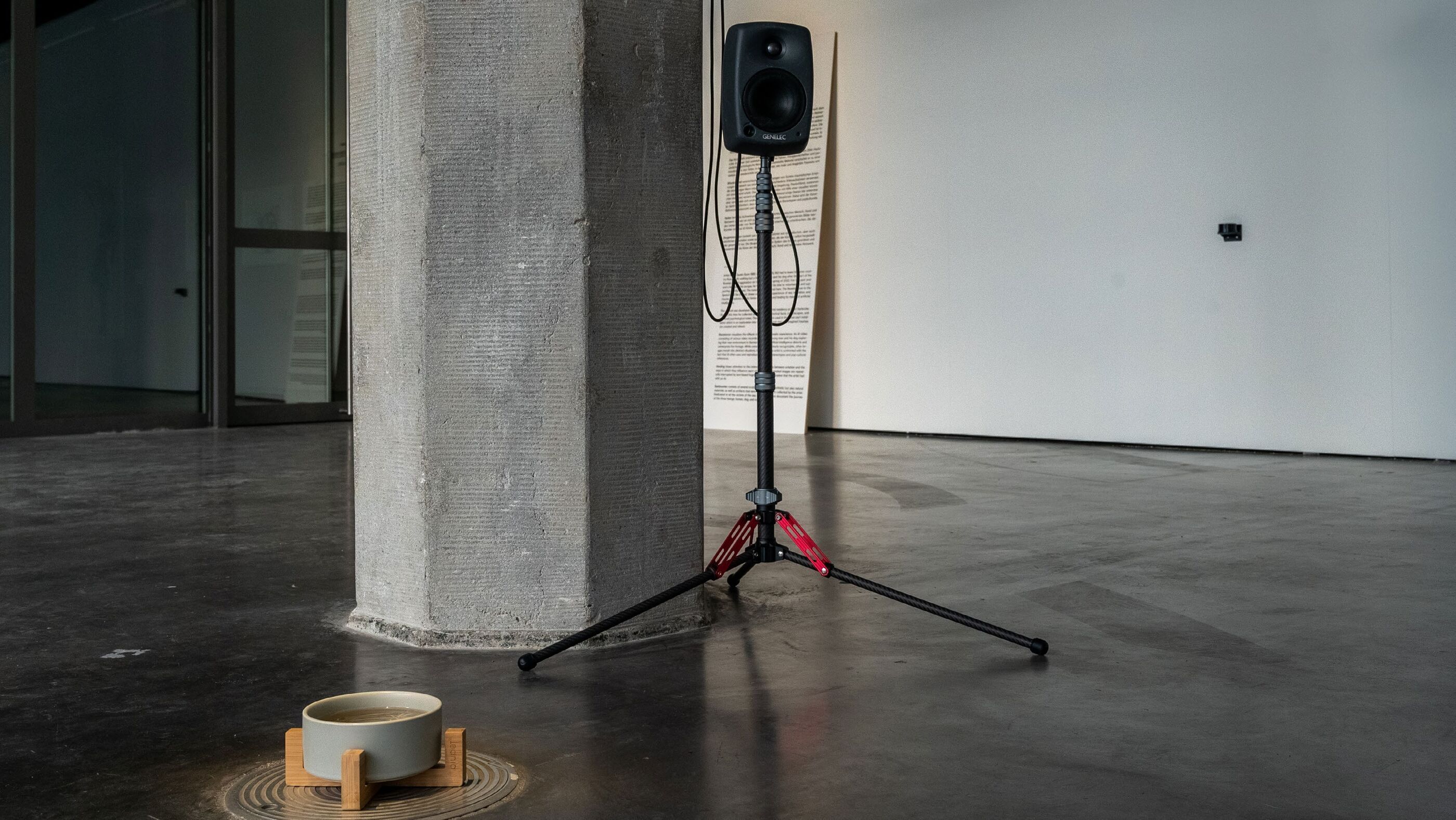 Small speaker on a tripod stand beside a concrete pillar, with a water bowl on a wooden stand and printed wall text behind