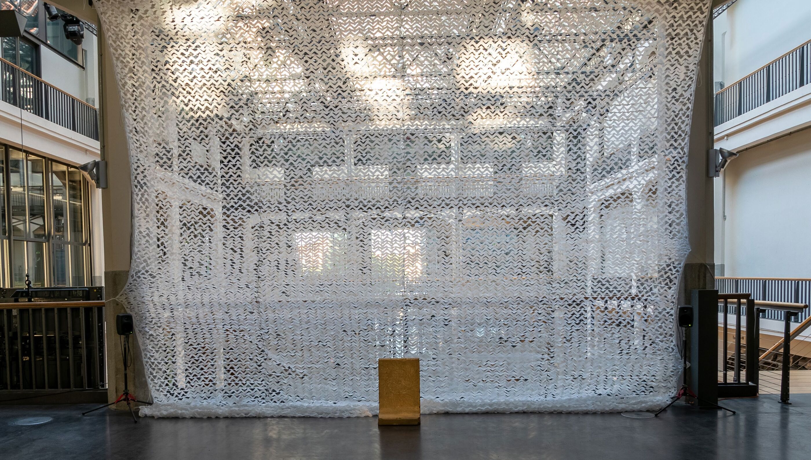 Gallery installation view of a large white patterned mesh curtain hanging between two levels of a stairwell, with a small block on the floor in front