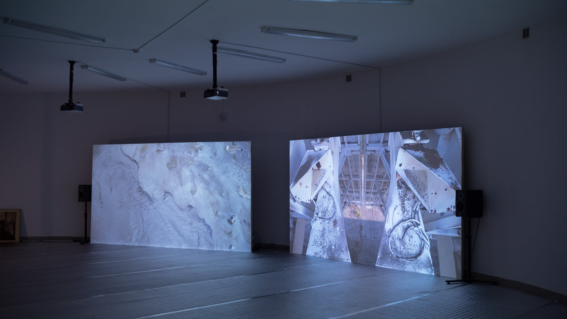 Gallery installation view of two large screens showing black-and-white photographs, one of a mineral surface and one of rusted machinery under a bridge, with a framed picture on the floor at left
