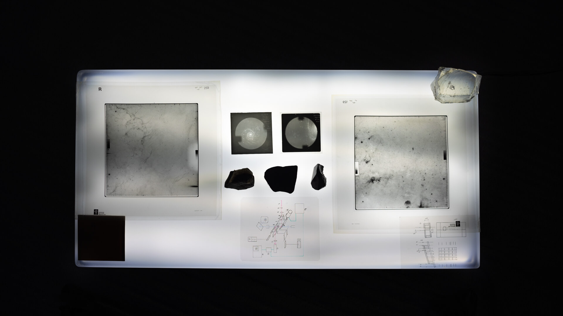 Overhead view of a large illuminated light table displaying photographic plates, star-chart negatives, dark mineral fragments, and technical diagrams