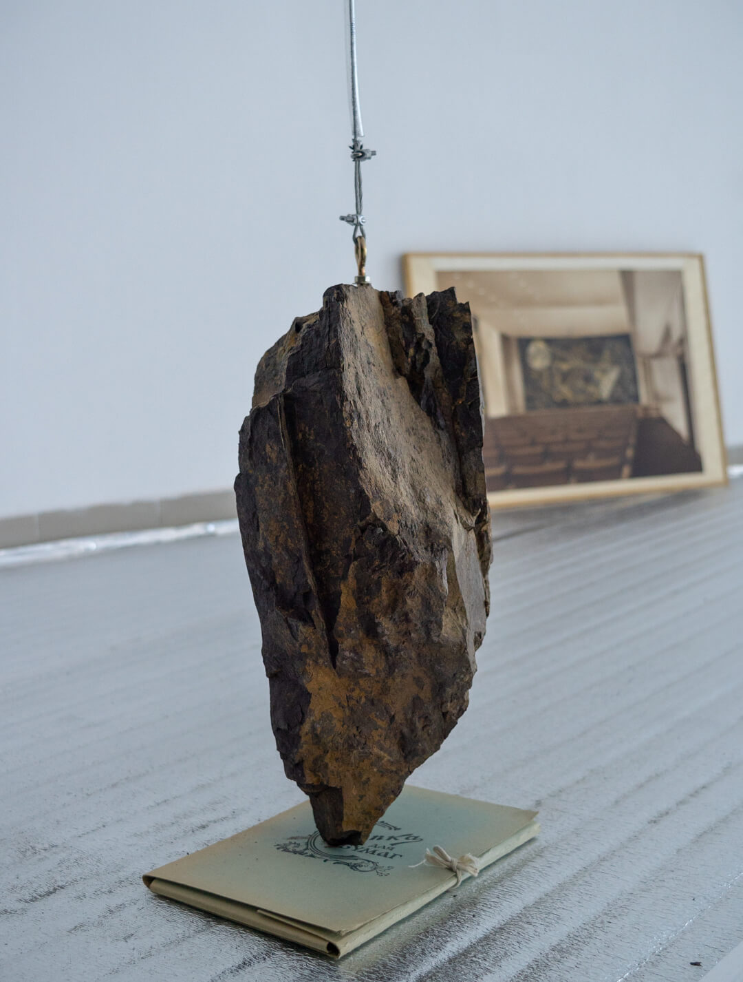 Jagged dark rock fragment suspended by a wire cable, hanging above a small tied notebook, with a framed photo of an auditorium in the background