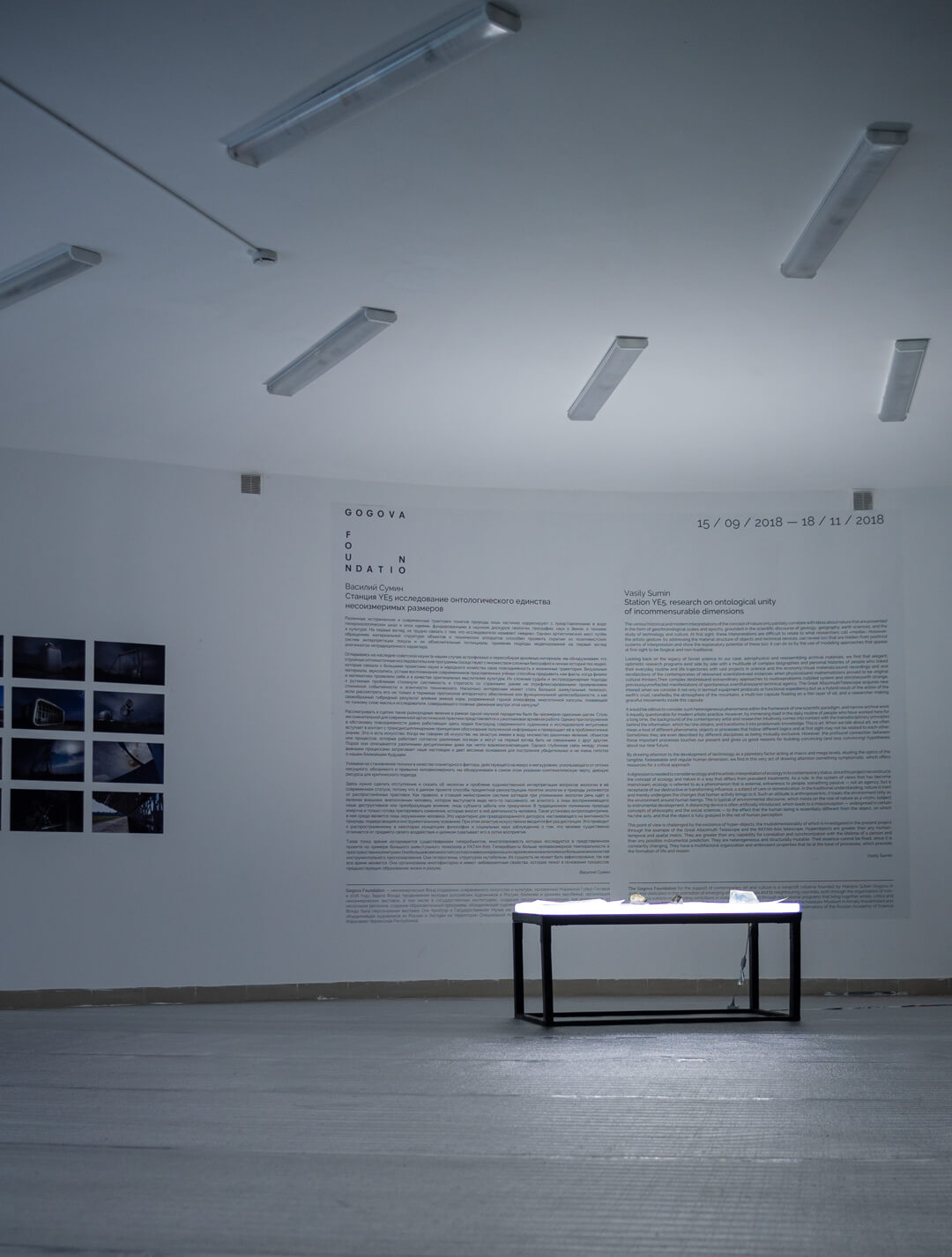 Gallery installation view of a wall text panel with framed black-and-white photographs beside an illuminated light table displaying objects