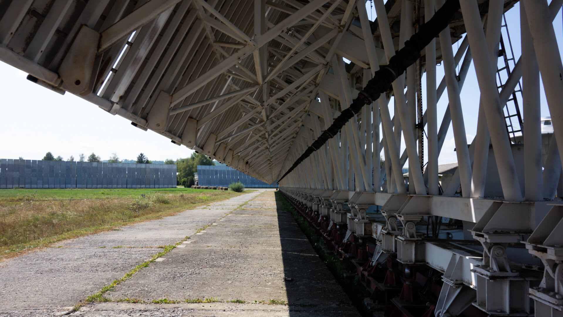 View along the underside of a long segmented radio telescope antenna structure, angled steel trusses stretching into the distance beside a concrete path