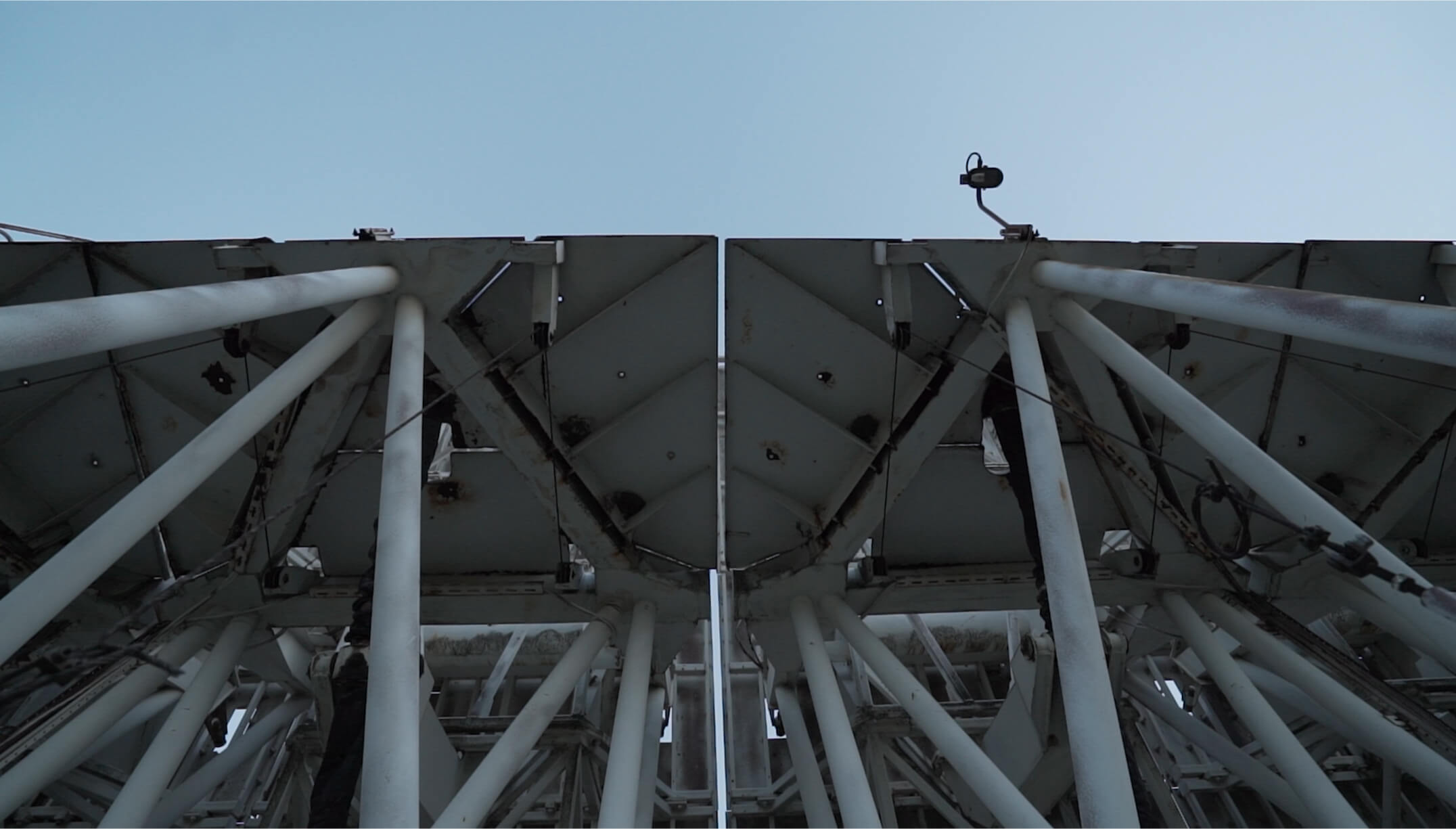Low-angle view looking straight up between two mirrored triangular sections of a telescope's white steel support structure against the sky