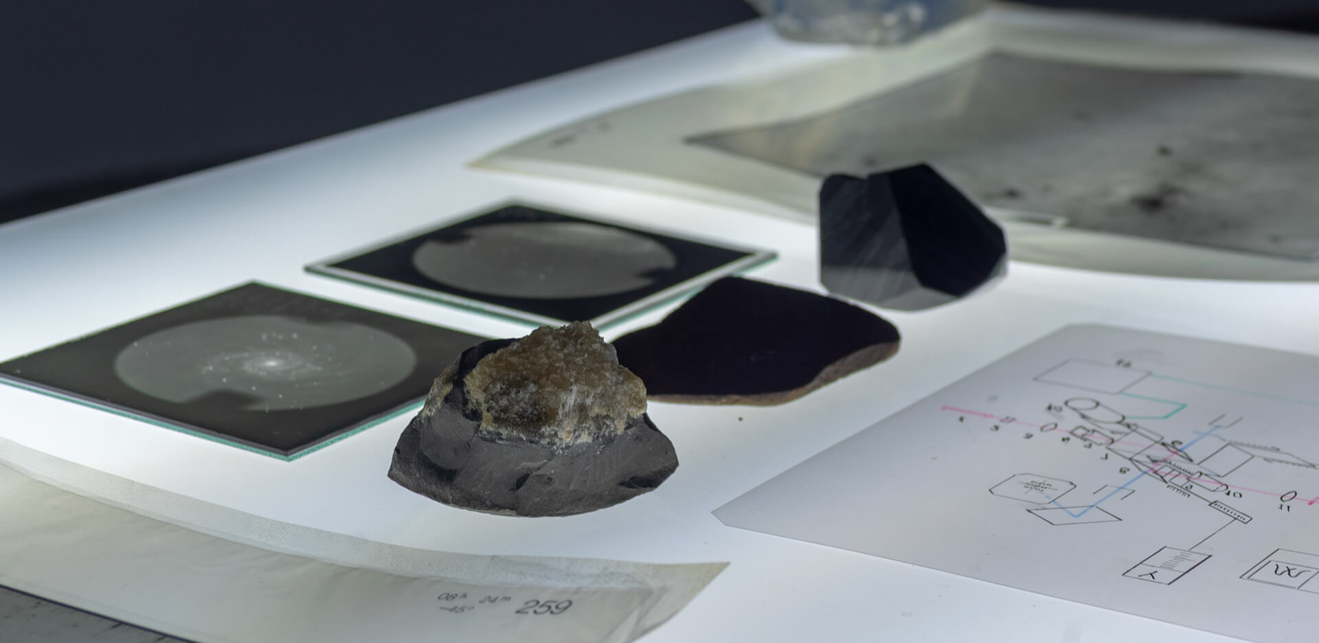 Close-up on a light table with rock fragments, glass photographic plates showing starfields, and a printed diagram, against a dark background